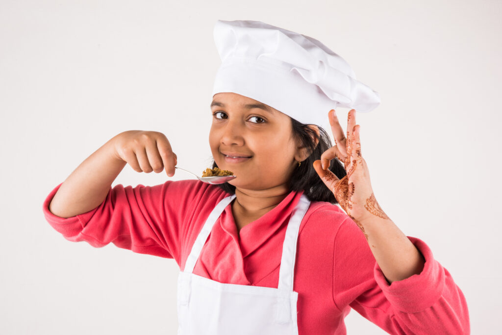 Cute little Indian kid or girl dressed as a Chef and holding cooking utensils or vegetables, standing isolated over white background